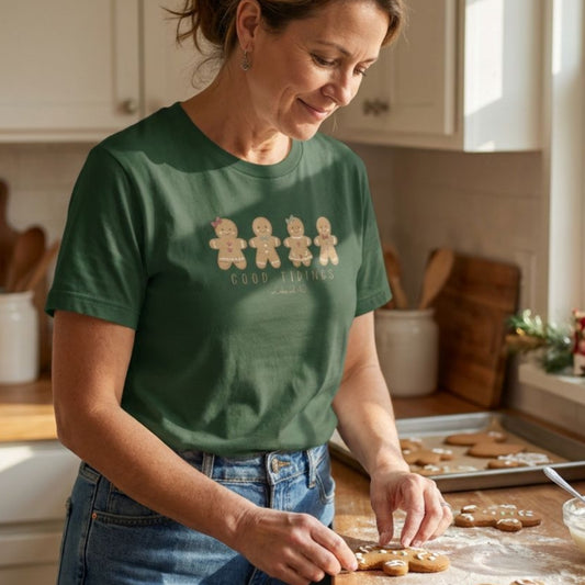 A woman in a green graphic t-shirt and blue jeans is shown in a warm, sunlit kitchen, decorating gingerbread cookies on a wooden counter. The t-shirt features a graphic of four gingerbread men with the words 'GOOD TRADITIONS' printed in gold.