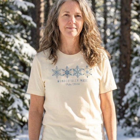 A woman with long wavy light brown hair stands outdoors in a snowy forest wearing a cream-colored t-shirt with a blue snowflake graphic and the text 'WONDERFULLY MADE