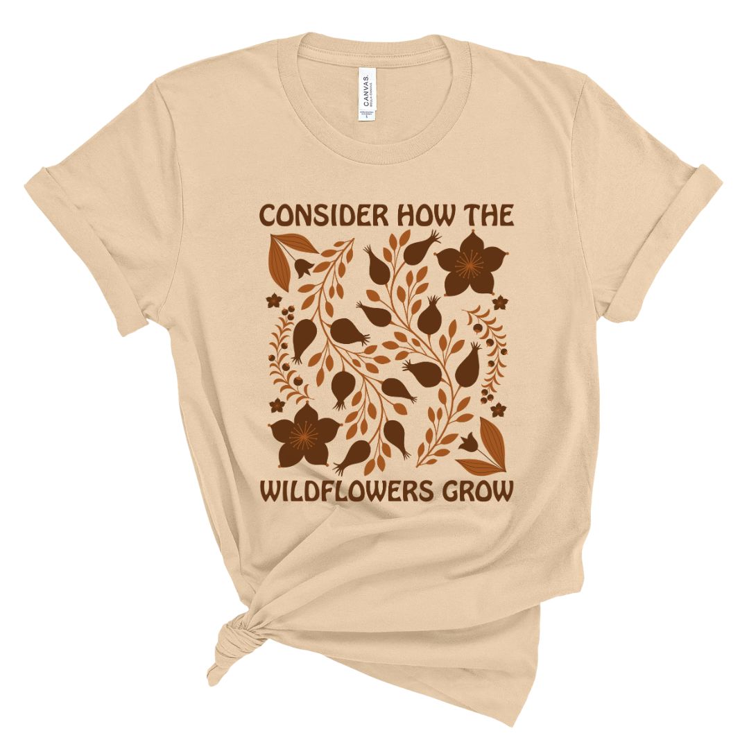 A beige colored t-shirt with a floral pattern and the text 'CONSIDER HOW THE WILDFLOWERS GROW' printed on the front.
