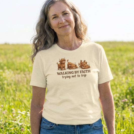 A woman standing in a grassy field wearing a cream-colored t-shirt with a graphic of three dogs and the text 'WALKING BY FAITH trying not to trip'.