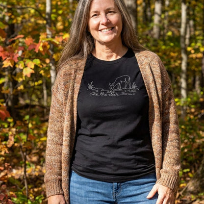 A woman standing outdoors in a wooded area with autumn foliage, wearing a black graphic T-shirt with a white deer design and the words 'as the deer,' layered under a brown knit cardigan and blue jeans.