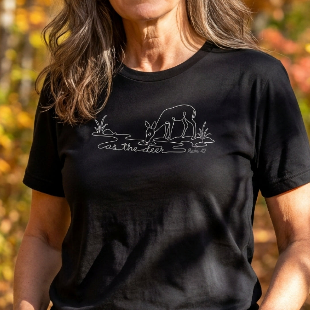 A woman wearing a black short-sleeve t-shirt with a white line art graphic of a deer drinking water and the text 'as the deer' and 'psalm 42'. The background is a blurred outdoor autumn scene.