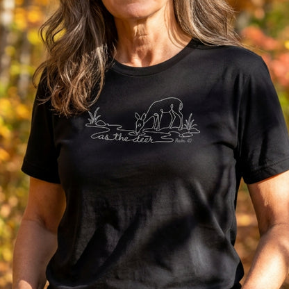 A woman wearing a black short-sleeve t-shirt with a white line art graphic of a deer drinking water and the text 'as the deer' and 'psalm 42'. The background is a blurred outdoor autumn scene.
