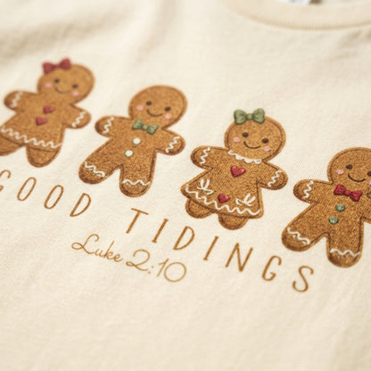 Close-up of a cream-colored garment featuring embroidered gingerbread men and the text 'GOOD TIDINGS Luke 2:10' in brown