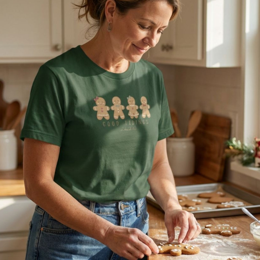A woman in a green graphic t-shirt and blue jeans is shown in a warm, sunlit kitchen, decorating gingerbread cookies on a wooden counter. The t-shirt features a graphic of four gingerbread men with the words 'GOOD TRADITIONS' printed in gold.