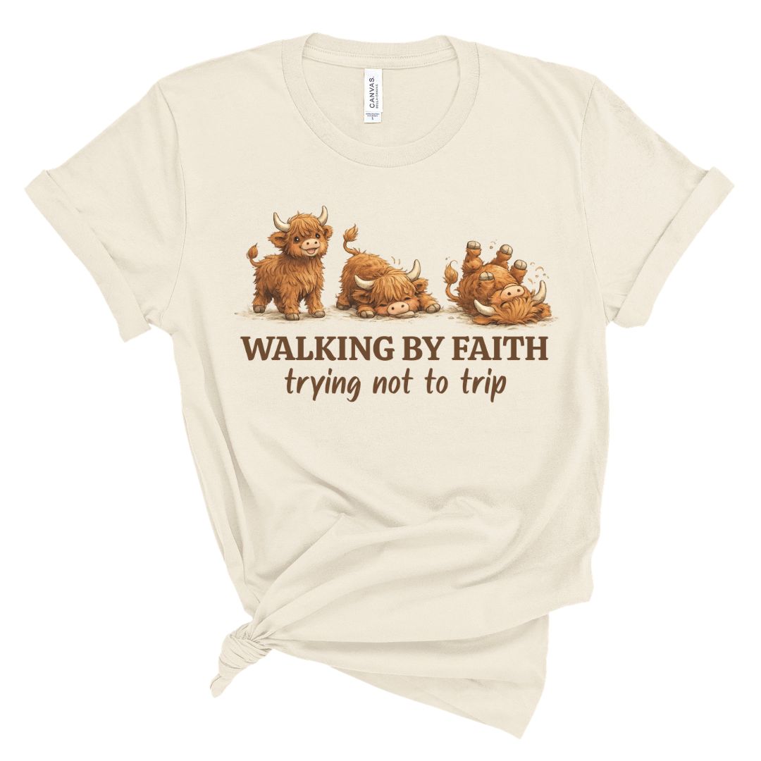 An off-white short-sleeve t-shirt with a graphic of four cute Highland cows and the text 'WALKING BY FAITH trying not to trip' printed on the front, styled with a knot at the bottom hem.