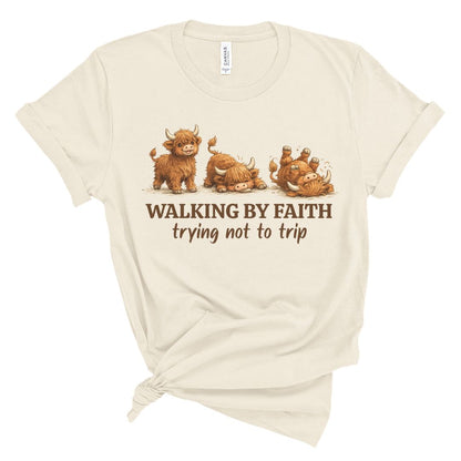 An off-white short-sleeve t-shirt with a graphic of four cute Highland cows and the text 'WALKING BY FAITH trying not to trip' printed on the front, styled with a knot at the bottom hem.