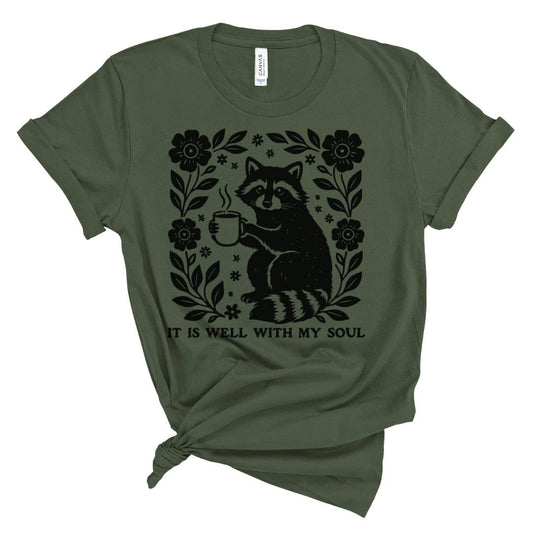 Christian t shirt for women with black woodland raccoon holding coffee and floral frame with It Is Well With My Soul text.