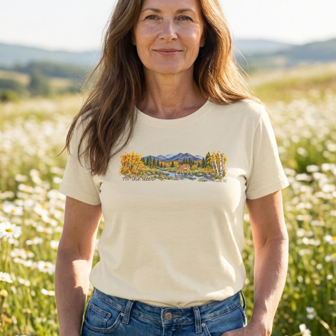 A woman wearing a cream-colored t-shirt with a colorful landscape graphic of mountains, trees, and a river, standing in a sunny field of white flowers.