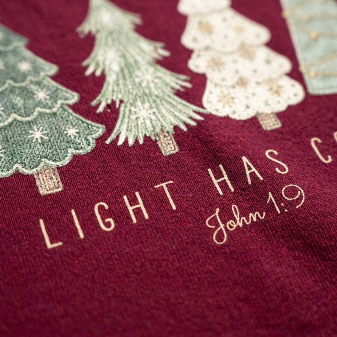 Close-up of a burgundy fabric surface with embroidered Christmas trees in green and cream and the words “LIGHT HAS COME” and “John 1:9”