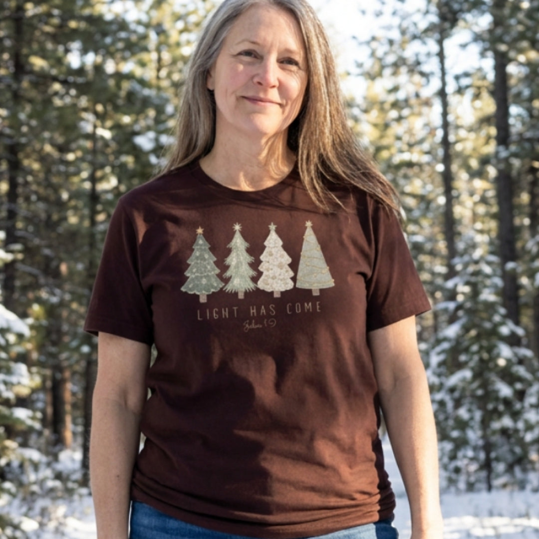 A woman wearing a brown short-sleeve t-shirt with a graphic of four Christmas trees and the text 'LIGHT HAS COME' standing in a snowy forest.