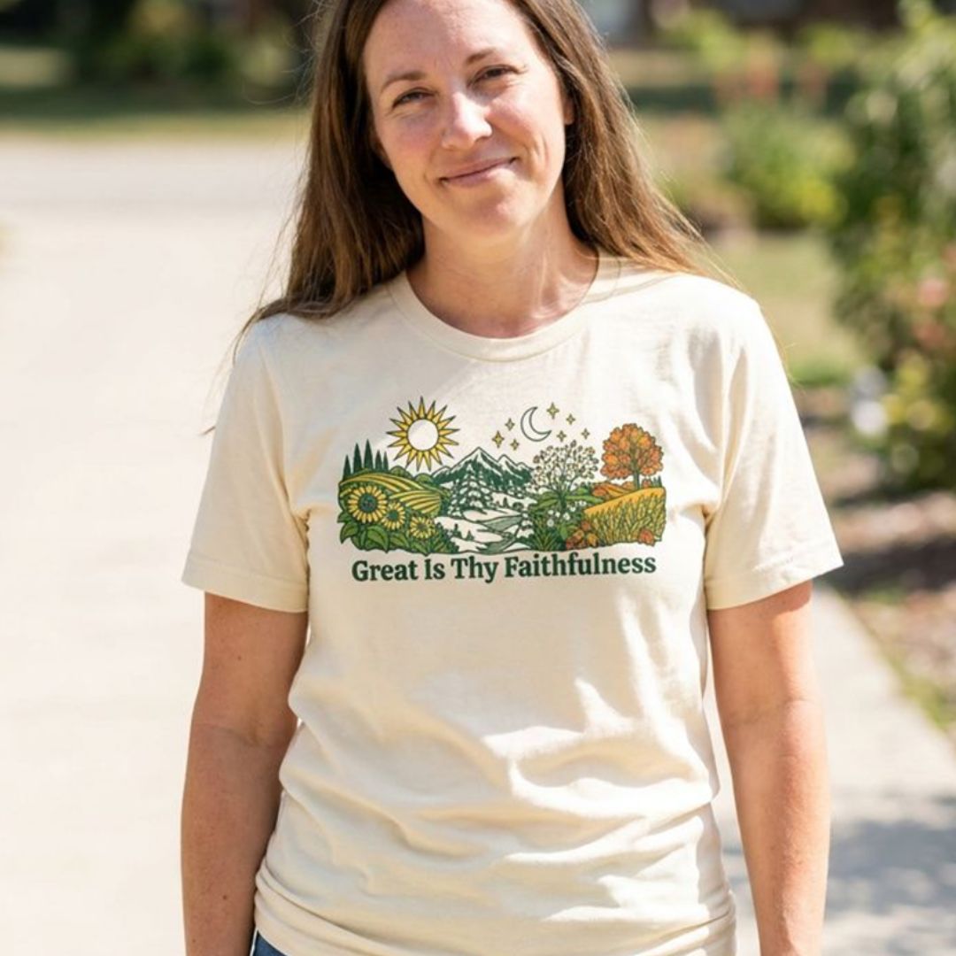 A woman wearing a cream-colored graphic t-shirt with a scenic landscape print and the text 'Great Is Thy Faithfulness' on a sunny outdoor path.