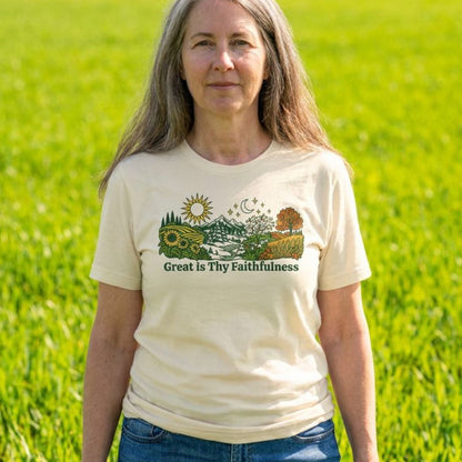 A woman standing in a grassy field wearing a cream-colored graphic t-shirt with a landscape illustration and the text 'Great Is Thy Faithfulness'.
