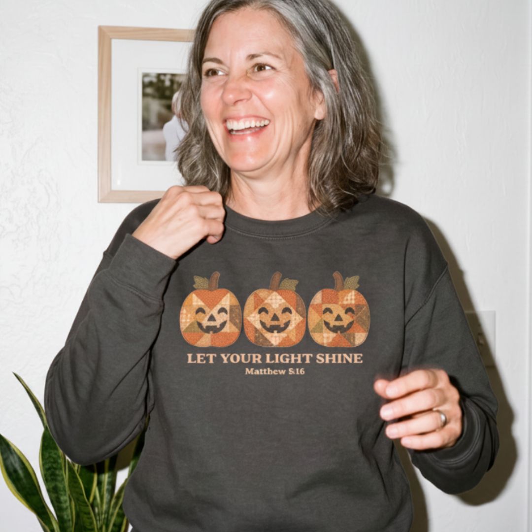 A woman smiling and wearing a dark charcoal gray long-sleeve crewneck sweatshirt with a graphic of three jack-o'-lanterns in a geometric pattern and the text 'LET YOUR LIGHT SHINE Matthew 5:16'.