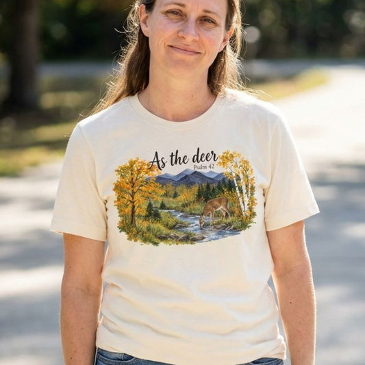 A woman wearing a cream-colored t-shirt with a graphic print of a deer in a forest stream, with the text 'As the deer Psalm 42' on the front, photographed outdoors on a sunny day.