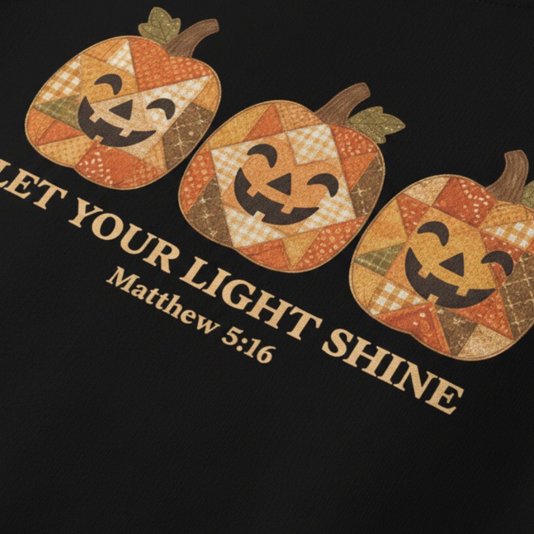 A close-up of a black garment featuring a graphic print of three patchwork-style jack-o'-lantern pumpkins and the text 'LET YOUR LIGHT SHINE' with 'Matthew 5:16' underneath.