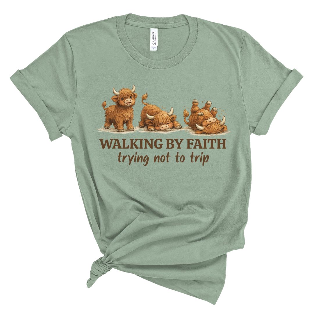 A sage green graphic t-shirt featuring three cartoon Highland cows and the text 'WALKING BY FAITH trying not to trip' in brown. The t-shirt is styled with a knot at the bottom left hem against a plain white background.