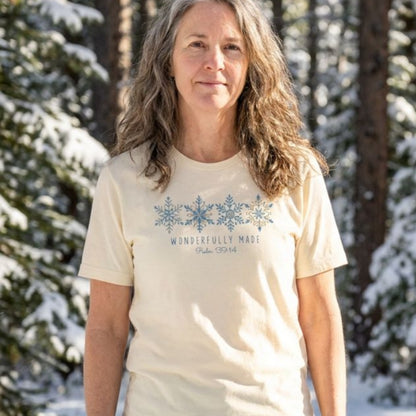 A woman with long wavy light brown hair stands outdoors in a snowy forest wearing a cream-colored t-shirt with a blue snowflake graphic and the text 'WONDERFULLY MADE