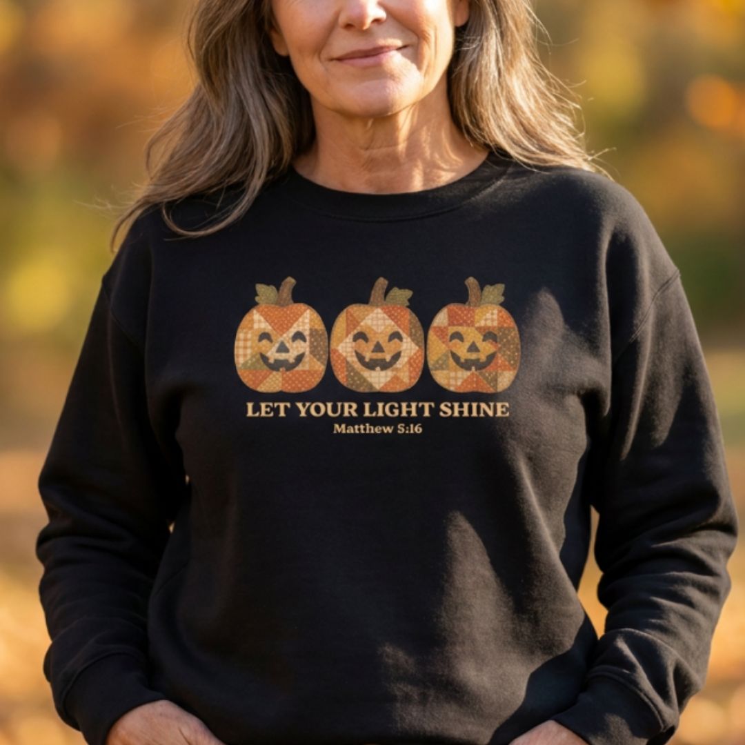 A woman wearing a black crewneck sweatshirt with a graphic of three patterned jack-o'-lanterns and the text 'LET YOUR LIGHT SHINE Matthew 5:16' on the front, set against a blurred autumn background.