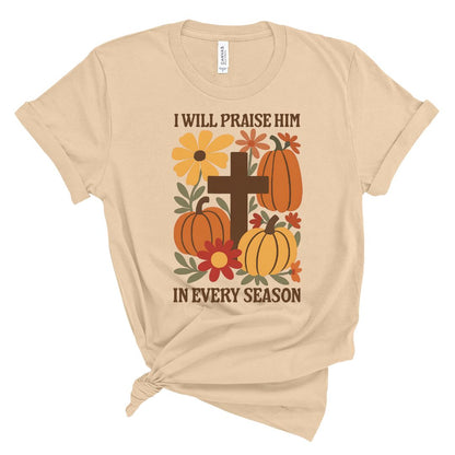Beige t-shirt with pumpkins, flowers, and cross design on a white background that says I will praise him in every season