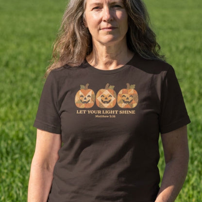 A woman wearing a dark brown t-shirt with a graphic of three jack-o'-lanterns and the text 'LET YOUR LIGHT SHINE Matthew 5:16' printed on the chest, set against a blurred green grass background.