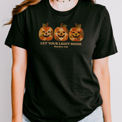 A woman wearing a black short-sleeve t-shirt with a graphic of three orange and brown plaid jack-o'-lanterns and the text 'LET YOUR LIGHT SHINE Matthew 5:16'.