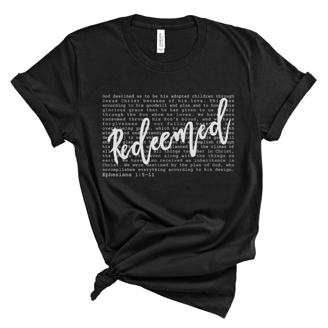 Black Christian women’s t-shirt with the word “Redeemed” in white script lettering layered over Ephesians 1:5–11 scripture text. Minimalist faith-based tee with a soft, relaxed fit.