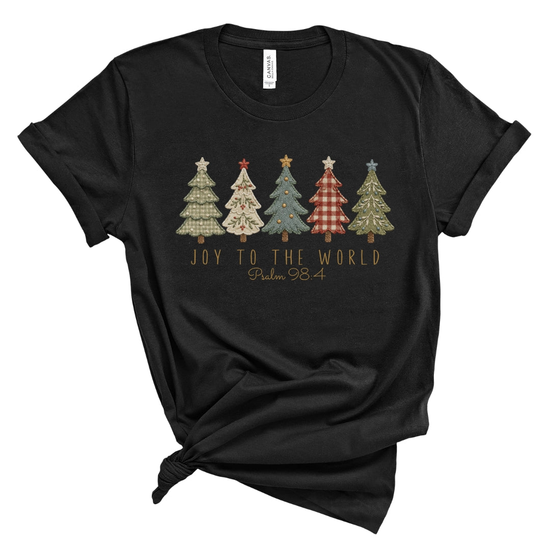 A black short-sleeve t-shirt with a graphic of five patterned Christmas trees and the text 'JOY TO THE WORLD Psalm 98:4' printed on the front, styled with a knot at the bottom hem on a white background.