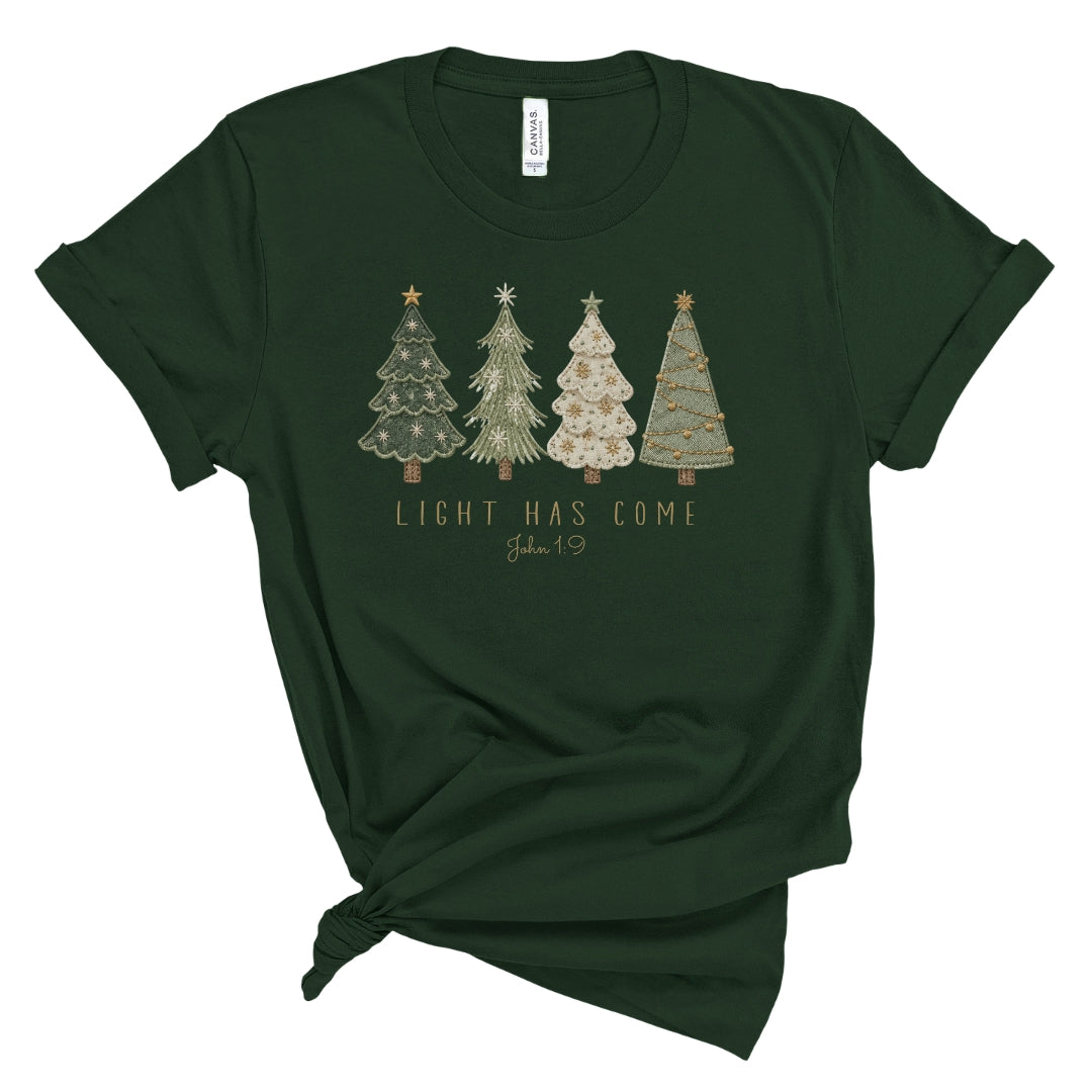 A dark forest green short-sleeve t-shirt with a knotted hem, featuring a graphic of four decorated Christmas trees and the text 'LIGHT HAS COME John 1:9' printed on the chest. The shirt is displayed flat against a solid white background.