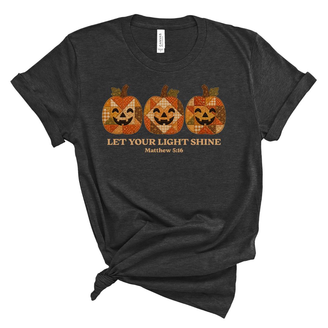 A dark heather gray t-shirt with a distressed graphic of three orange and brown plaid jack-o'-lanterns above the text 'LET YOUR LIGHT SHINE Matthew 5:16'. The t-shirt is styled with a knot at the bottom hem and is photographed on a solid white background.