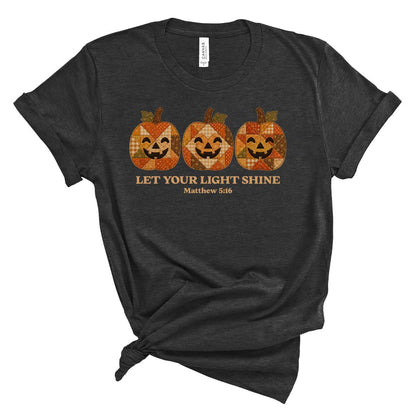 A dark heather gray t-shirt with a distressed graphic of three orange and brown plaid jack-o'-lanterns above the text 'LET YOUR LIGHT SHINE Matthew 5:16'. The t-shirt is styled with a knot at the bottom hem and is photographed on a solid white background.