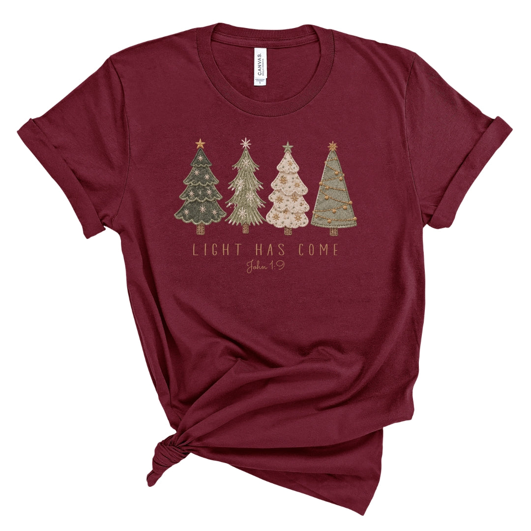A maroon short-sleeve t-shirt with a knotted hem, featuring a graphic of four decorated Christmas trees and the text 'LIGHT HAS COME John 1:9' printed on the front. The shirt is displayed flat against a white background.