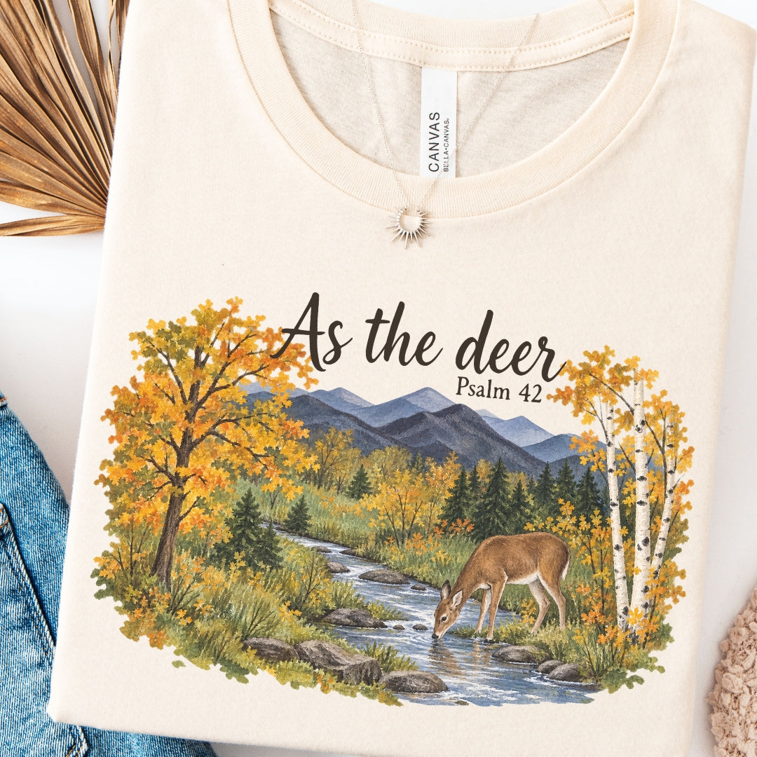 A cream-colored short-sleeve t-shirt with a graphic print of a deer drinking from a stream in an autumn forest, with the text 'As the deer Psalm 42'. The t-shirt is displayed flat with styled props including a denim jacket, dried palm leaves, and a knit sweater.