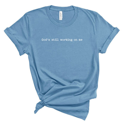 Christian t shirt for women with the phrase “God’s still working on me” in simple white lettering, minimalist faith based shirt for everyday wear.