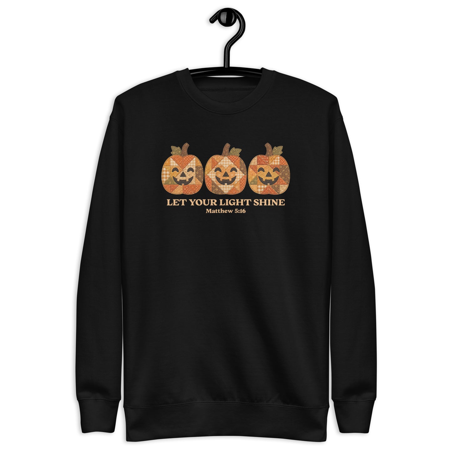 A black crewneck sweatshirt hanging on a black hanger against a plain white background. The sweatshirt features a graphic of three orange jack-o'-lantern pumpkins with a distressed, patterned texture, above the text 'LET YOUR LIGHT SHINE' and 'Matthew 5:16' in a gold color.