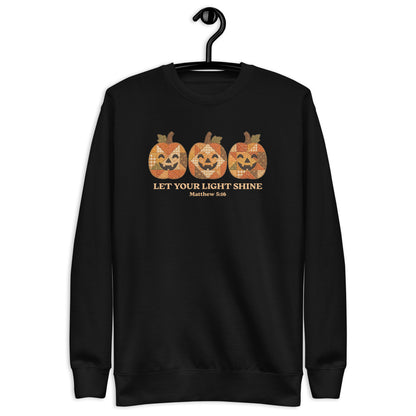 A black crewneck sweatshirt hanging on a black hanger against a plain white background. The sweatshirt features a graphic of three orange jack-o'-lantern pumpkins with a distressed, patterned texture, above the text 'LET YOUR LIGHT SHINE' and 'Matthew 5:16' in a gold color.