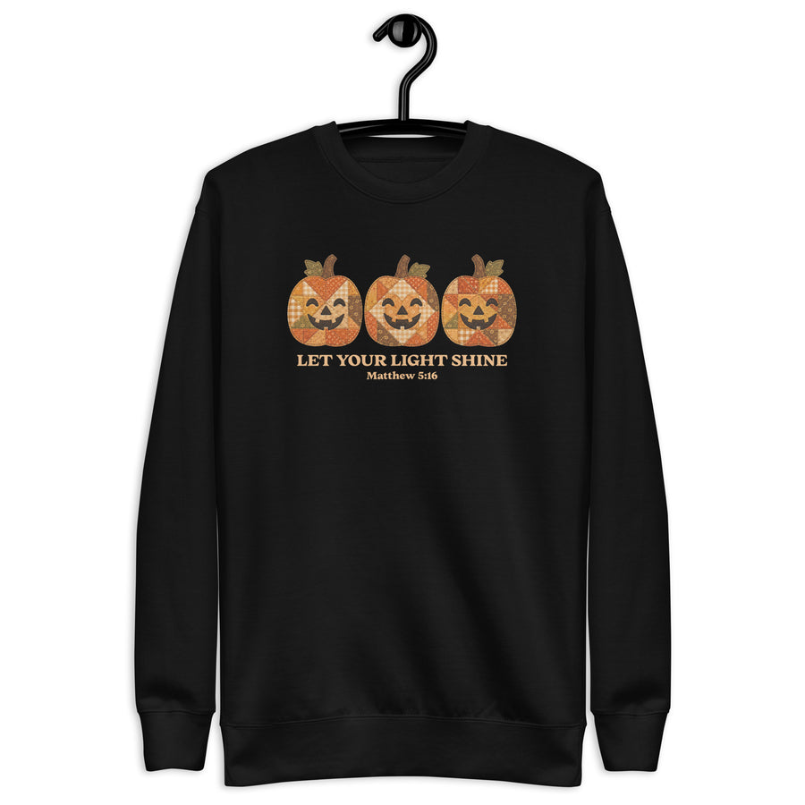 A black crewneck sweatshirt hanging on a black hanger against a plain white background. The sweatshirt features a graphic of three orange jack-o'-lantern pumpkins with a distressed, patterned texture, above the text 'LET YOUR LIGHT SHINE' and 'Matthew 5:16' in a gold color.