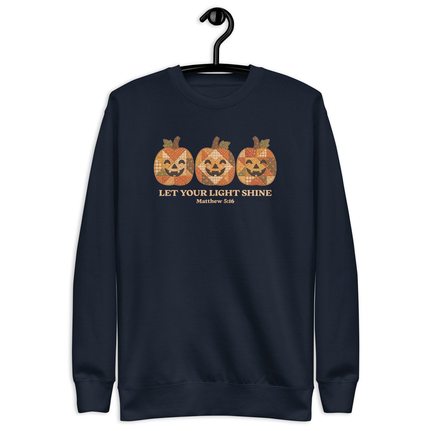 crewneck sweatshirt hanging on a black hanger against a plain white background. The sweatshirt features a graphic of three orange jack-o'-lantern pumpkins with a distressed, patterned texture, above the text 'LET YOUR LIGHT SHINE' and 'Matthew 5:16' in a gold color