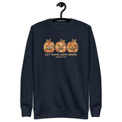 crewneck sweatshirt hanging on a black hanger against a plain white background. The sweatshirt features a graphic of three orange jack-o'-lantern pumpkins with a distressed, patterned texture, above the text 'LET YOUR LIGHT SHINE' and 'Matthew 5:16' in a gold color