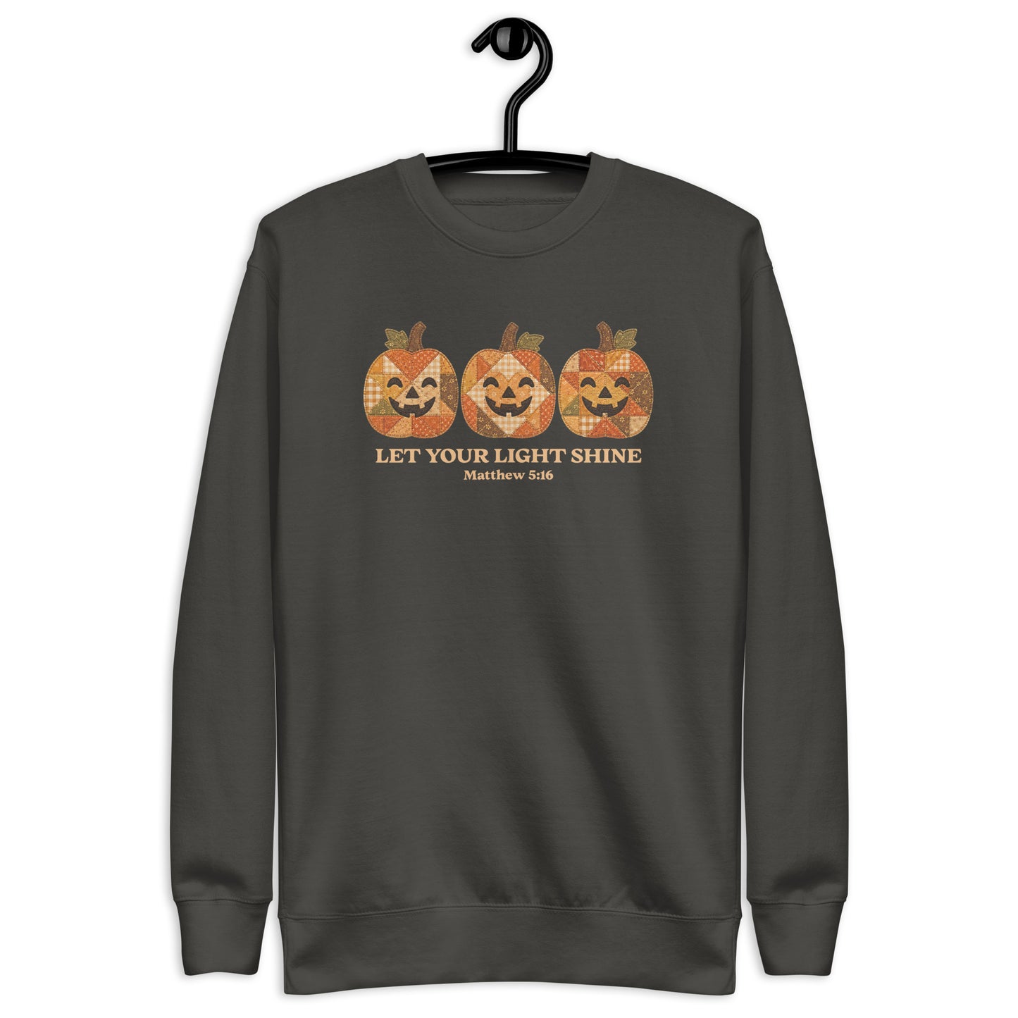 crewneck sweatshirt hanging on a black hanger against a plain white background. The sweatshirt features a graphic of three orange jack-o'-lantern pumpkins with a distressed, patterned texture, above the text 'LET YOUR LIGHT SHINE' and 'Matthew 5:16' in a gold color.