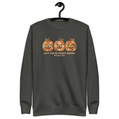 crewneck sweatshirt hanging on a black hanger against a plain white background. The sweatshirt features a graphic of three orange jack-o'-lantern pumpkins with a distressed, patterned texture, above the text 'LET YOUR LIGHT SHINE' and 'Matthew 5:16' in a gold color.