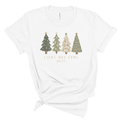 A white short-sleeve t-shirt with a knotted hem, featuring a graphic of four decorated Christmas trees and the text 'LIGHT HAS COME 2025' printed on the front. The shirt is displayed flat against a white background.