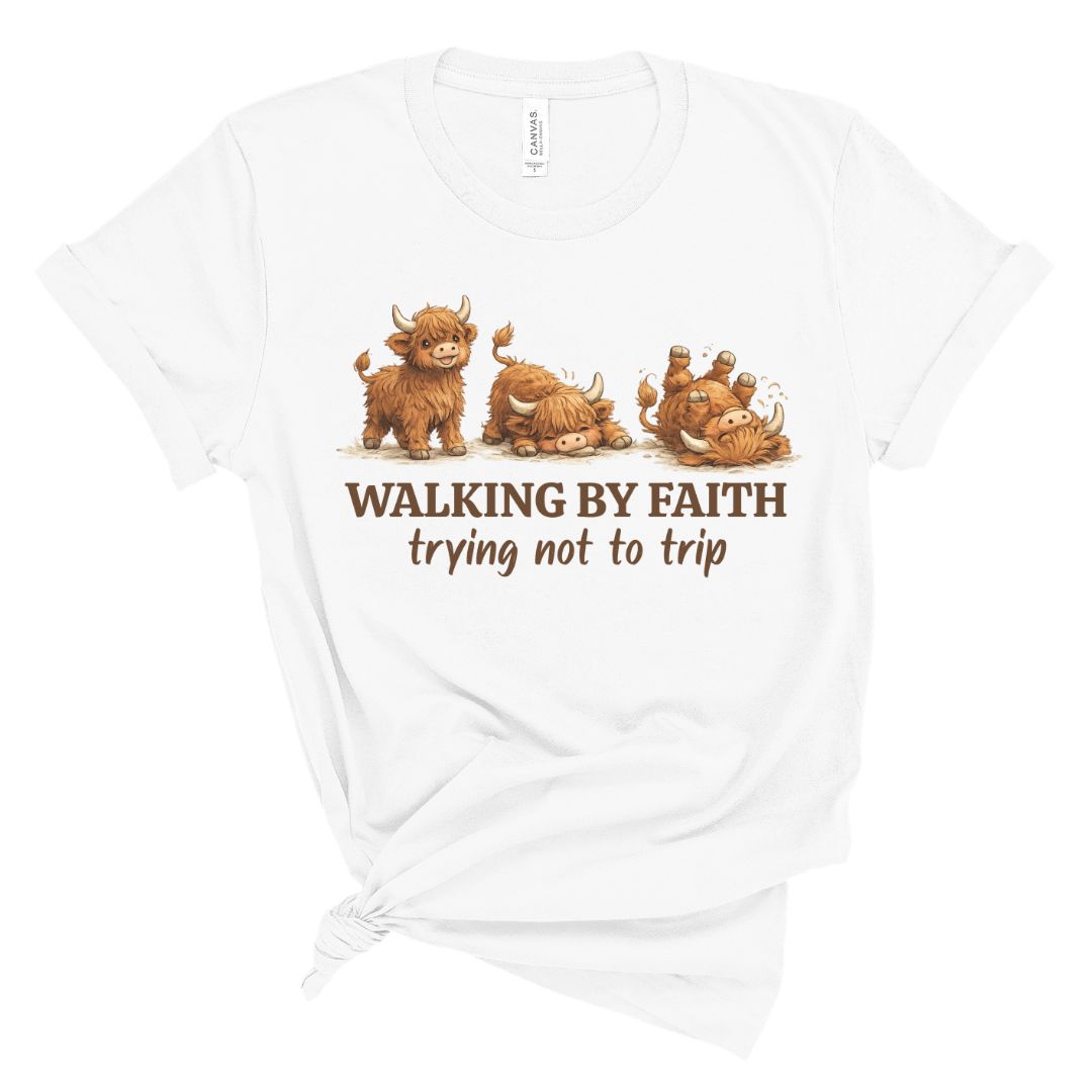 A white short-sleeve t-shirt with a graphic of four brown highland cows on a white background. Below the cows, the text reads 'WALKING BY FAITH trying not to trip' in brown.